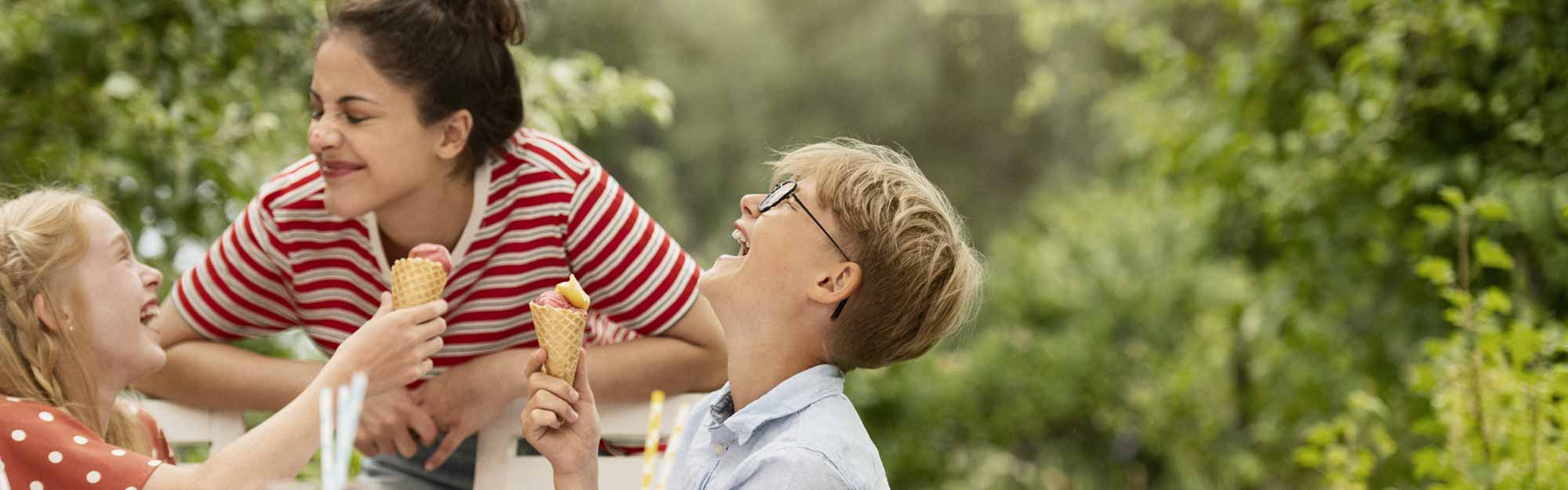 Två barn och en kvinna skrattar och äter glasstrutar i trädgården
